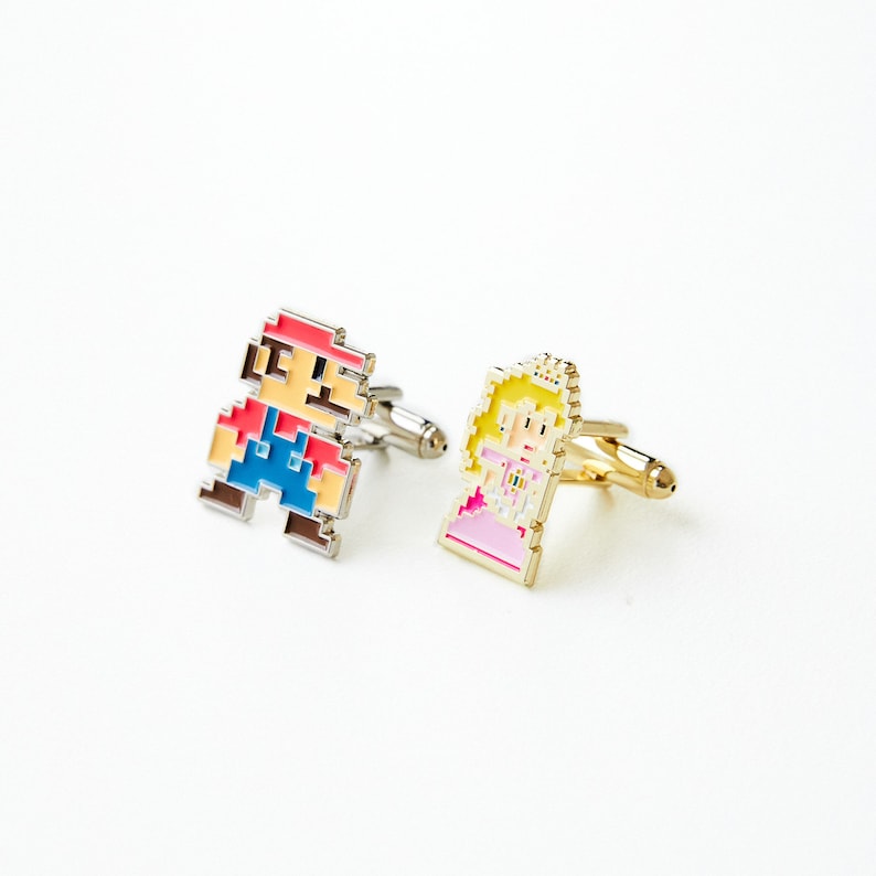 May include: A pair of cufflinks featuring pixelated characters from a popular video game. One cufflink features a red and blue character with a mustache, while the other cufflink features a pink and yellow character with a crown.