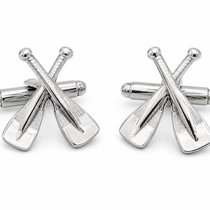 May include: A pair of silver-toned cufflinks featuring a design of two crossed oars. Each cufflink has a polished metallic finish with textured detailing on the blades of the oars and a standard toggle closure mechanism on the back for secure fastening.