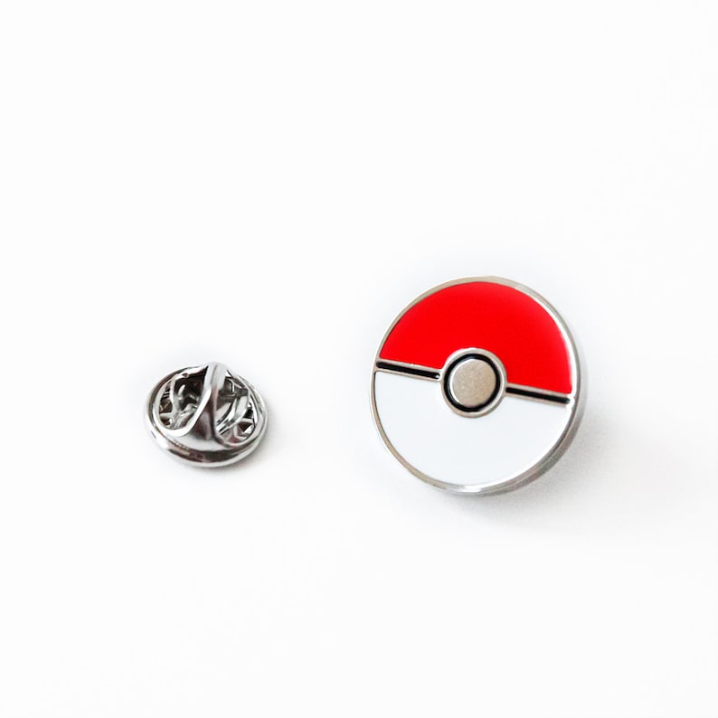 May include: A silver metal pin with a red and white Pokeball design. The pin has a silver metal clasp on the back.