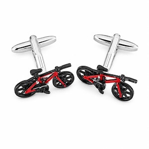 Könnte beinhalten: Ein Paar silberfarbene Manschettenknöpfe mit einem Miniatur-Fahrrad-Design. Jedes Fahrrad ist mit einem leuchtend roten Rahmen, schwarzen Rädern, einem schwarzen Lenker und einem schwarzen Sattel bemalt. Die Manschettenknöpfe haben einen Standard-Knebelverschluss.