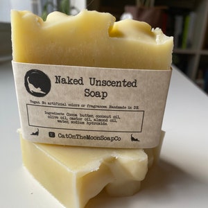May include: Two bars of naked unscented soap, stacked on top of each other. The top bar has a brown paper label that reads "Naked Unscented Soap" and lists the ingredients: Cocoa butter, coconut oil, olive oil, castor oil, almond oil, water, and sodium hydroxide. The label also states that the soap is vegan, contains no artificial colours or fragrances, and is handmade in Germany. The label includes the company name "CatOnTheMoonSoapCo".