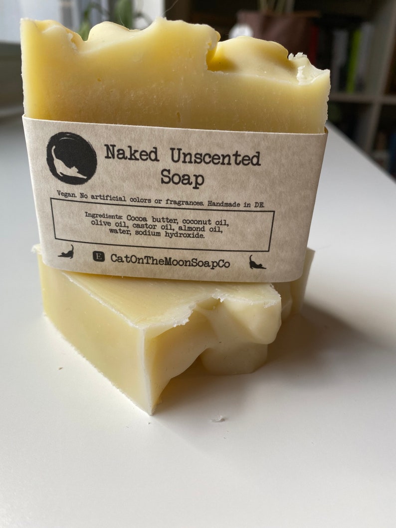 May include: Two bars of naked unscented soap, stacked on top of each other. The top bar has a brown paper label that reads "Naked Unscented Soap" and lists the ingredients: Cocoa butter, coconut oil, olive oil, castor oil, almond oil, water, and sodium hydroxide. The label also states that the soap is vegan, contains no artificial colours or fragrances, and is handmade in Germany. The label includes the company name "CatOnTheMoonSoapCo".