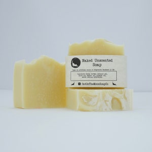 May include: Three bars of unscented, natural soap. The soap is a light yellow colour and is stacked on top of each other. The top bar has a label that reads "Naked Unscented Soap" and lists the ingredients: Cocoa butter, coconut oil, olive oil, castor oil, almond oil, water, and sodium hydroxide. The label also includes the text "Vegan. No artificial colours or fragrances. Handmade in the UK." and the website address "CatOnTheMoonSoapCo."