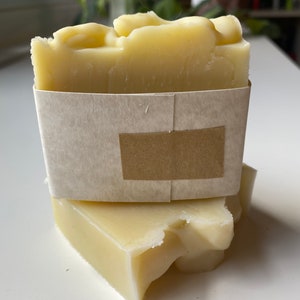 May include: Two bars of handmade soap wrapped in brown paper. The soap is a pale yellow colour and has a rough texture.