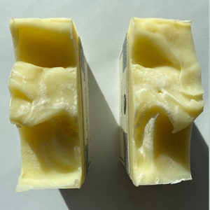 May include: Two bars of pale yellow soap, each with a white label on the side. The soap is slightly textured and appears to be handmade.