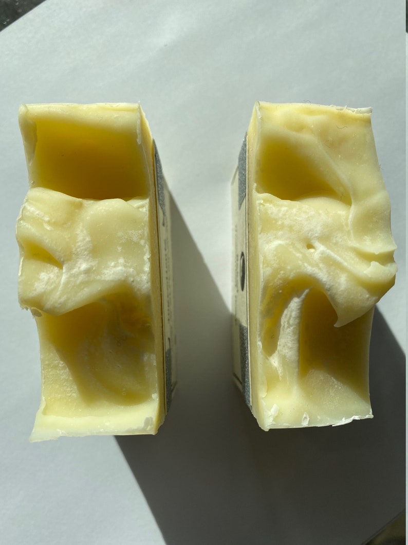 May include: Two bars of pale yellow soap, each with a white label on the side. The soap is slightly textured and appears to be handmade.