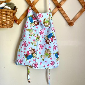 Peut inclure: Un tablier bleu clair avec un motif de cœurs roses et de poupées tenant des ours en peluche. Le tablier a deux longues attaches.