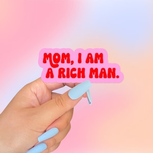 May include: Pink sticker with the text "MOM, I AM A RICH MAN." in red, bubbly letters. The sticker is being held by a hand with light blue nail polish.