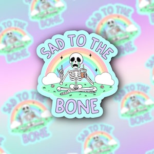 May include: A sticker with a cartoon skeleton sitting under a rainbow with a cup of coffee. The text reads "Sad to the Bone".