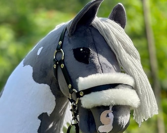 May include: A soft toy hobby horse in a gray and white spotted design. It has a black bridle with silver-tone fittings and a white, fluffy noseband. The horse has a white mane and a black lead rope, with a blurred green backdrop.