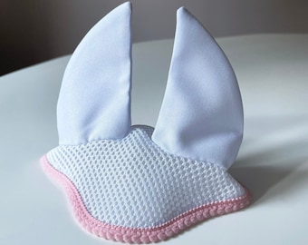 Könnte beinhalten: Weißer Fliegenhaube für Pferde mit rosa Besatz. Die Haube ist aus Netzstoff und hat zwei große Ohren.