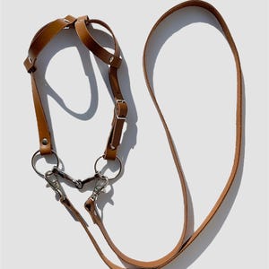 May include: A brown leather camera strap with two metal clips and a long strap. The strap has a ring on each side and a buckle in the middle.
