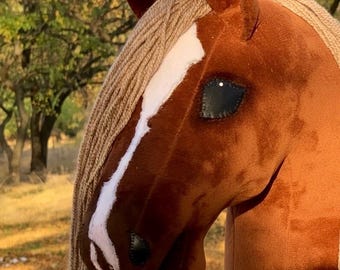 May include: A plush brown horse head with a white stripe down the face and a tan mane. The horse has a black eye and is set against a blurred background of trees and foliage. This toy is suitable for children.