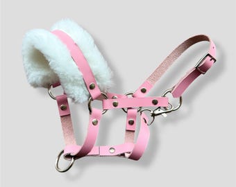 Puede incluir: Un cabestro de cuero rosa con acolchado de piel sintética blanca. El cabestro tiene herrajes metálicos plateados y un cierre de presión.