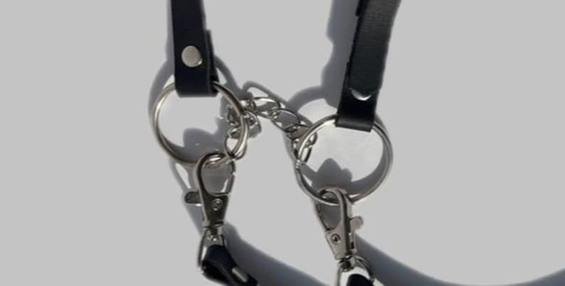 May include: Close-up of a black leather strap with two silver key rings and a silver chain connecting them.