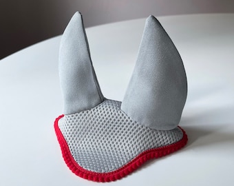 Könnte beinhalten: Eine graue Fliegenhaube für Pferde mit zwei aufrechten Ohrenschützern und einem Netzmittelteil. Die Haube ist mit einem roten Rand versehen.