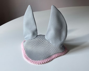 Könnte beinhalten: Ein grauer Fliegenhaube für Pferde mit rosa Besatz. Die Haube hat Netzohren und einen weichen, gepolsterten Rand.