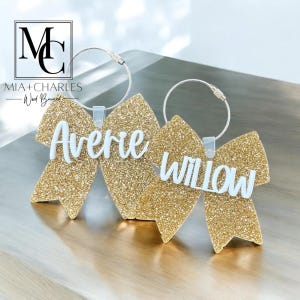 May include: Two glittery gold bow-shaped luggage tags with white lettering spelling "Avere" and "Willow". Each tag has a silver metal loop for attachment. The tags are on a wooden surface. The top left corner has the text "MIA+CHARLES - Wood Burned".