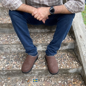 May include: A person wearing brown wool slippers with a black rubber sole. The slippers are sitting on a set of concrete steps. The person is wearing blue jeans and a plaid shirt. The text "KLÄGN SWEDISH COMFORT" is visible at the bottom of the image.