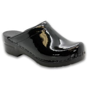 May include: A black, glossy clog shoe with a rounded toe and a low heel. The shoe has a smooth, patent leather-like finish and is stitched around the sole. The shoe is designed for comfort and support.