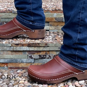 May include: Brown leather clogs with a wooden sole and a white stitch detail. The clogs are worn by a person wearing blue jeans. The person is standing on a stone staircase.