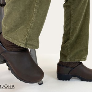 May include: A pair of dark brown clogs with a smooth finish and black trim around the collar. The clogs feature a thick, black, molded sole and a rounded toe box. The brand name BJÖRK SWEDISH COMFORT is printed in the bottom left corner with a flag.
