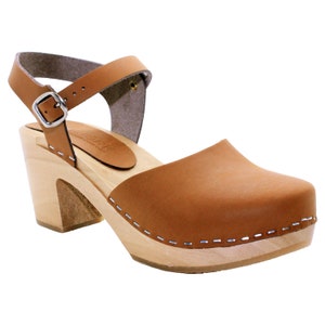 May include: Tan leather heeled clog with an ankle strap and buckle. The clog has a wooden sole and a stitched edge.
