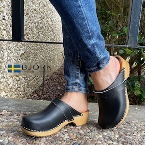 May include: Black leather clogs with a wooden sole and a strap across the top. The clogs are worn with blue jeans. The brand name "BJÖRK SWEDISH COMFORT" is visible in the image.