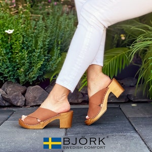 May include: Brown leather clogs with a criss-cross strap design and a wooden heel. The clogs are worn by a person wearing white jeans. The brand name "BJÖRK SWEDISH COMFORT" is displayed on the bottom of the image.
