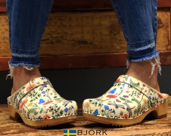 May include: A pair of cream-colored clogs with a floral and dragonfly print. The clogs have a wooden base and a leather strap.