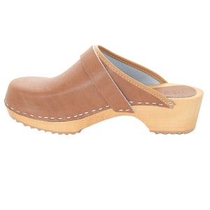 May include: Brown leather clog with a white stitched seam around the sole and a white stitched strap across the top. The clog has a wooden heel and a wooden base with a textured sole.
