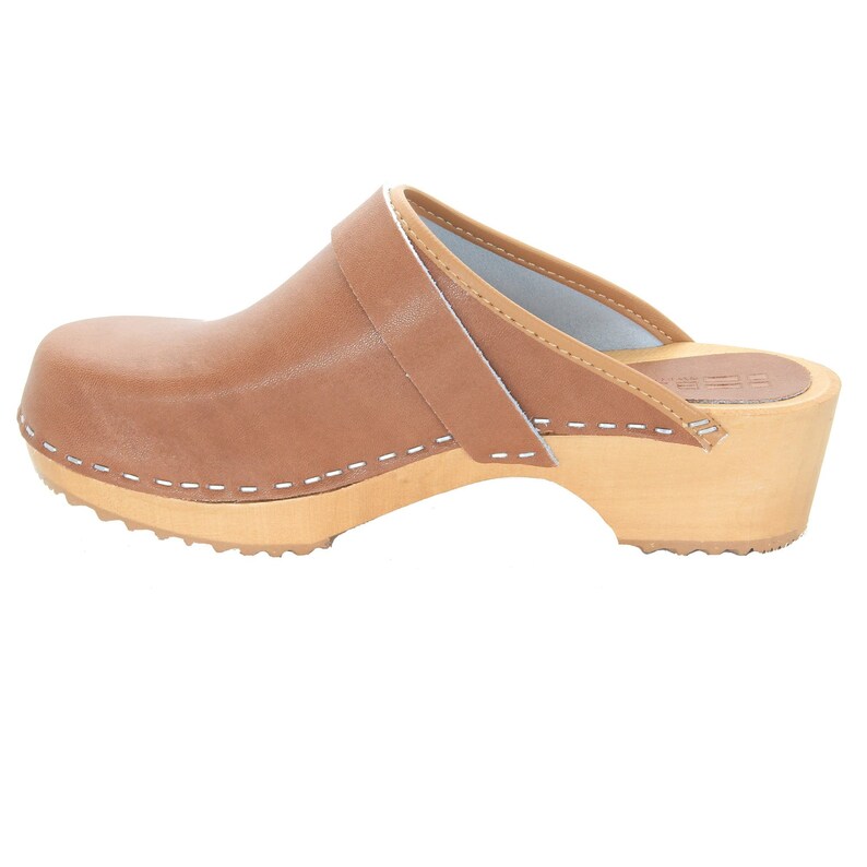 May include: Brown leather clog with a white stitched seam around the sole and a white stitched strap across the top. The clog has a wooden heel and a wooden base with a textured sole.