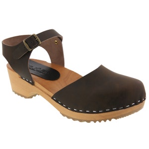 May include: Brown leather clog with an ankle strap and a wooden heel. The clog has a white stitch detail around the edge of the sole.