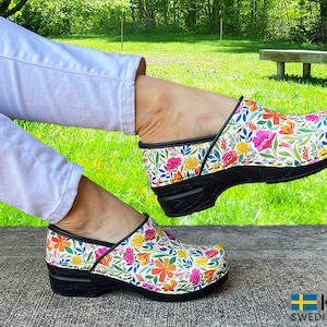 May include: Floral patterned clogs with a black sole and trim. The clogs feature a colorful design of pink, orange, yellow, and green flowers on a white background. The brand name "KLÄGN SWEDISH COMFORT" is visible.
