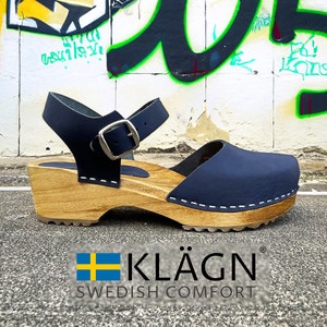 May include: Dark blue leather clogs with a wooden platform sole and an adjustable ankle strap. The clogs have a white stitching detail around the sole and a silver buckle on the strap. The brand name "KLÄGN" is printed on the sole, along with the text "SWEDISH COMFORT".