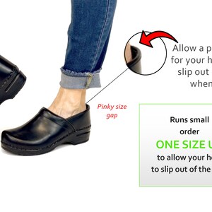 Op de afbeelding: Zwarte leren klompen met een houten hak. De afbeelding toont een persoon die de klompen draagt en een notitie dat de schoenen klein uitvallen en dat je een maat groter moet bestellen om te zorgen dat de hiel uit de klomp kan glijden tijdens het lopen.