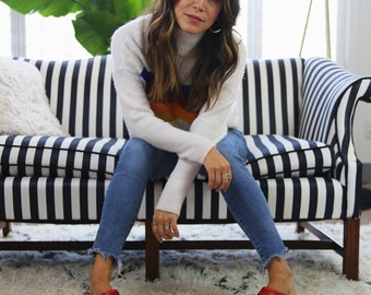 May include: A woman wearing a white sweater with a rainbow design, blue jeans, and red clogs. She is sitting on a black and white striped sofa. The rug is white with a black geometric pattern.