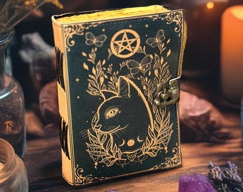 May include: A dark green journal with a cat design, pentagram, and botanical accents. The journal has a clasp closure and gold-colored edges. Amethyst and quartz crystals, candles, and dried flowers surround the journal on a wooden surface.