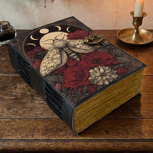 May include: A decorative book with a moth and floral design. The cover features a moth illustration with a moon and floral pattern in red, white, and black. The book has gold-colored edges and a black spine. The book is on a wooden surface.