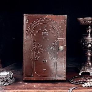 Puede incluir: Un diario encuadernado en cuero marrón con un diseño intrincado de árboles y estrellas. El diario tiene un cierre y está estampado con el texto "No todo lo que reluce es oro, no todos los que deambulan están perdidos; lo viejo que es fuerte no se marchita, las raíces profundas no son alcanzadas por la helada. De las cenizas se despertará un fuego, una luz de las sombras surgirá; renovada será la hoja que se rompió, el sin corona volverá a ser rey."