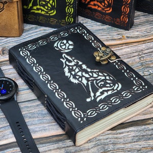 May include: A black leather journal with a wolf design and a metal clasp. The journal has a Celtic knot border and is closed. A black smartwatch with a round face is on a wooden surface.