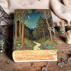 May include: A hardback journal with a forest scene illustration. The cover depicts a moonlit forest with a path, trees, and flowers. The journal has a metal clasp and a decorative bookmark.