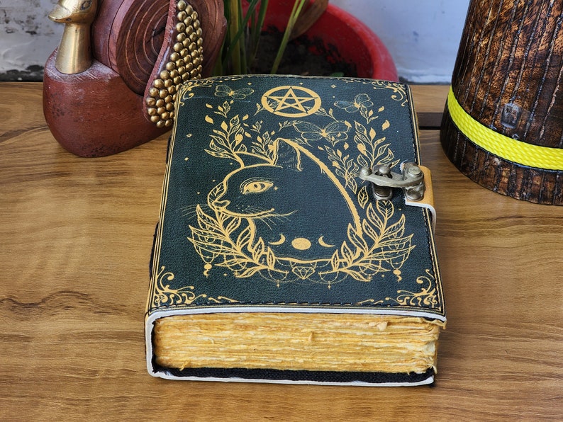 May include: A dark green journal with a cat design and a pentagram. The cover features a gold illustration of a cat's face, leaves, and a crescent moon. The journal has a metal clasp and aged-looking pages.