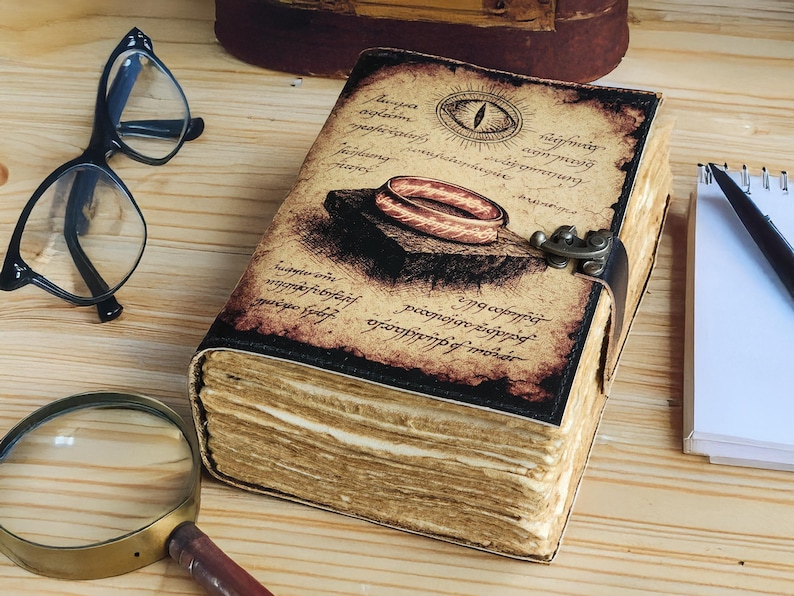 May include: A vintage-style journal with a detailed cover illustration featuring an eye and a ring. The aged pages are visible, and the journal is secured with a metal clasp. Also present are eyeglasses, a magnifying glass, and a pen.