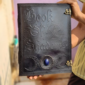 May include: A black leather journal with the words "Book of Shadows" embossed on the cover. The cover also features a pentagram, three crescent moons, and a blue gemstone.