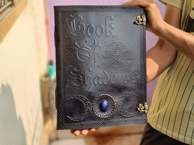 May include: A black leather journal with the words "Book of Shadows" embossed on the cover. The cover also features a pentagram, three crescent moons, and a blue gemstone.