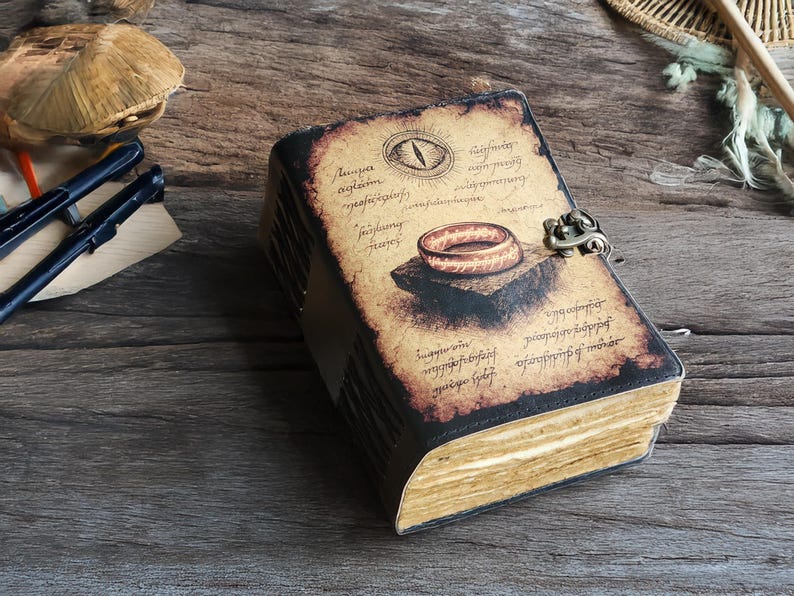 May include: A vintage-style journal with a leather cover and clasp. The cover features an illustration of a ring and eye, with aged paper and handwritten text. The journal is open, revealing aged pages, and rests on a wooden surface.