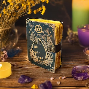 May include: A dark teal journal with a cat illustration, pentagram, and botanical accents. The pages have a golden hue, and the journal is secured with a black strap and clasp. The scene includes candles, crystals, and dried flowers.