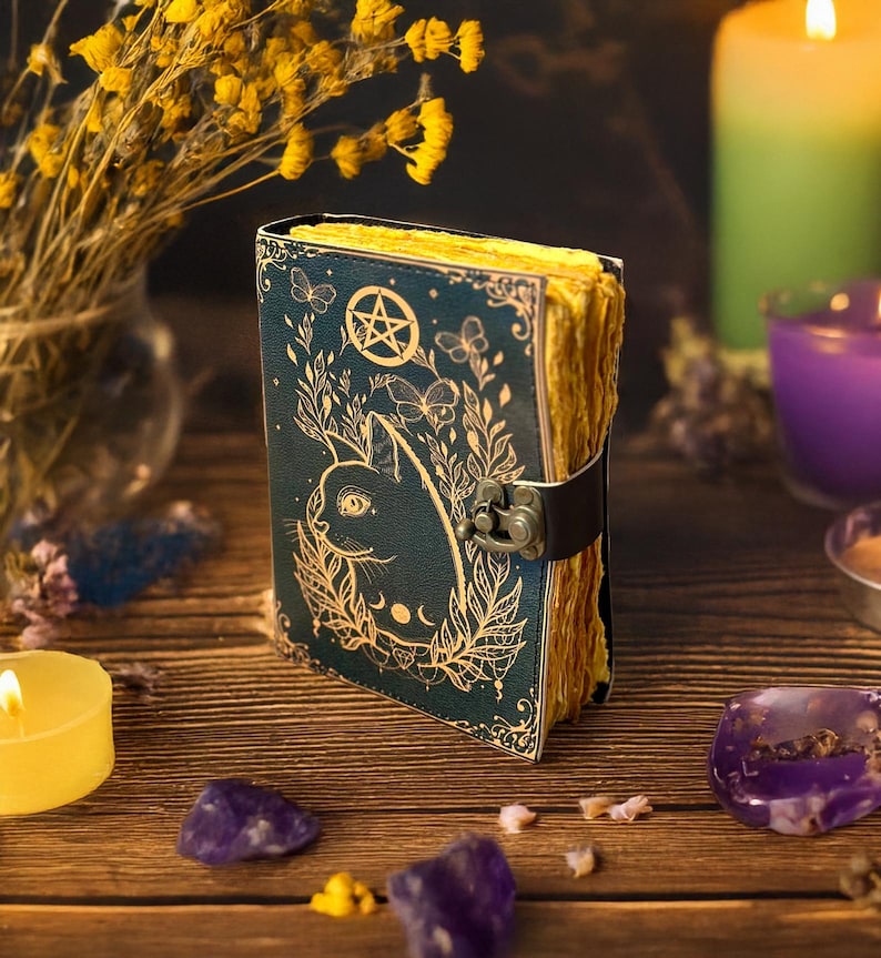 May include: A dark teal journal with a cat illustration, pentagram, and botanical accents. The pages have a golden hue, and the journal is secured with a black strap and clasp. The scene includes candles, crystals, and dried flowers.