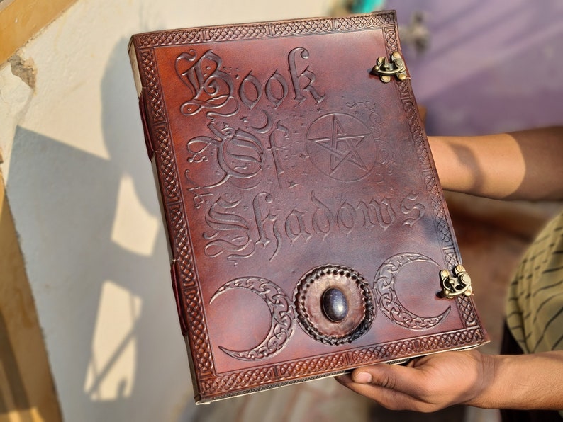 May include: A brown leather-bound journal with the words "Book of Shadows" embossed on the cover. The cover also features a pentagram, three crescent moons, and decorative metal clasps.